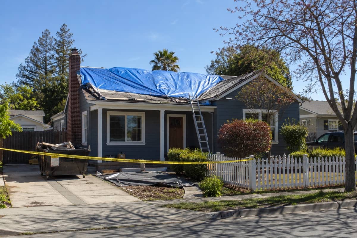 A house with a tarped, damaged roof, a dumpster in the driveway, and caution tape out front.